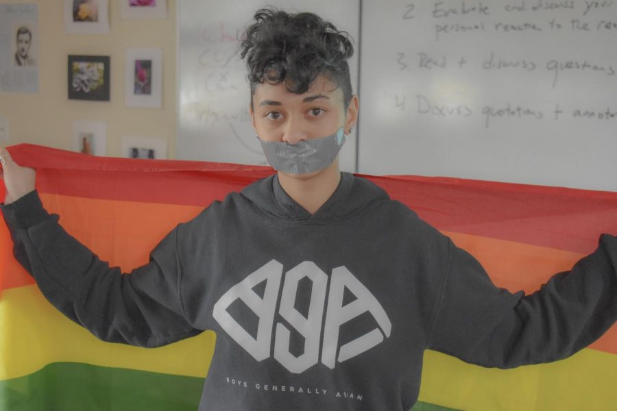 The leader of Woodsides Gay Straight Alliance holds a rainbow flag behind them,  duct tape over their mouth to signify hate speech.