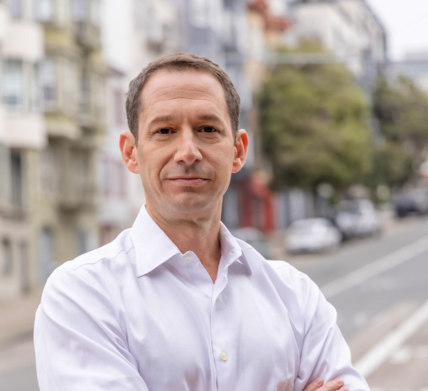 A headshot of Mayor Daniel Lurie with a blurred background of a steep San Francisco intersection